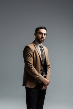 Şık giyinmiş, gözlüklü genç bir Arap gri kameraya bakıyor.