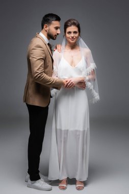 Gelinlik içinde mutlu bir gelin, kameraya bakıyor gri üzerinde Müslüman damadın yanında.