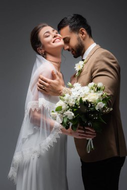 Müslüman damadın yanında düğün buketi tutan neşeli kadın gri yolda izole edilmiş.