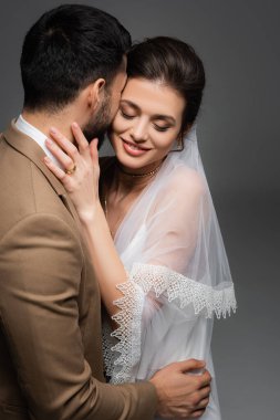 Güler yüzlü, gözleri kapalı, Müslüman nişanlısına sarılan gri elbiseli gelin.