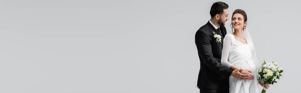 Улыбающийся мусульманский жених касается живота беременной невесты букетом, изолированным на сером, баннере 