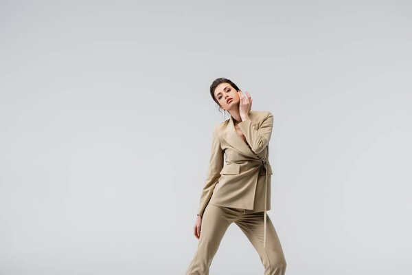 brunette woman in stylish pantsuit touching face while posing isolated on grey