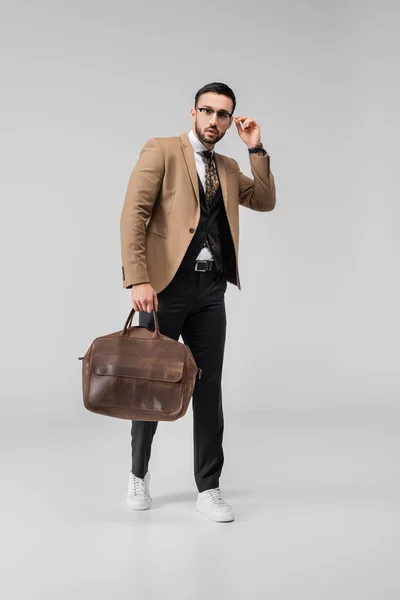 full length view of trendy arabian man touching eyeglasses while posing with leather bag on grey