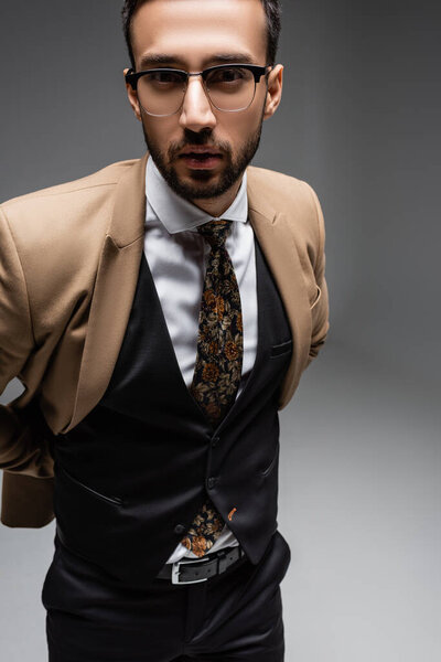 muslim man in beige blazer and black vest looking at camera on grey