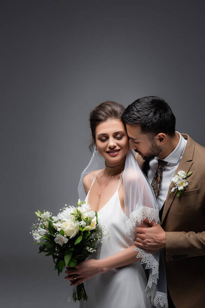 arabian man embracing happy bride holding wedding bouquet on grey