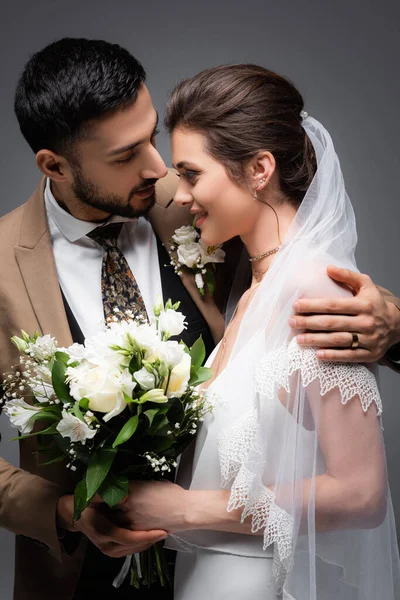 Zarif Arap damat, mutlu geline sarılıyor ve gri renkte izole edilmiş düğün buketi tutuyor.