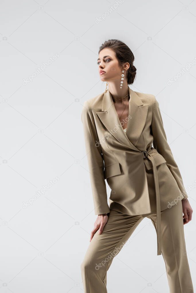 elegant woman in beige pantsuit looking away while standing isolated on grey