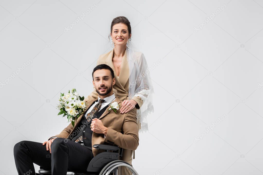 elegant woman in veil, with wedding bouquet, looking at camera near handicapped muslim fiance isolated on grey