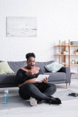 šokovaný africký Američan plus velikost žena ve sportovním oblečení pomocí digitální tablet v obývacím pokoji