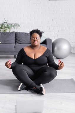 Afro-amerikan artı spor giyim bölümünde oturan kadın yoga pozu veriyor oturma odasında dizüstü bilgisayarın yanında.