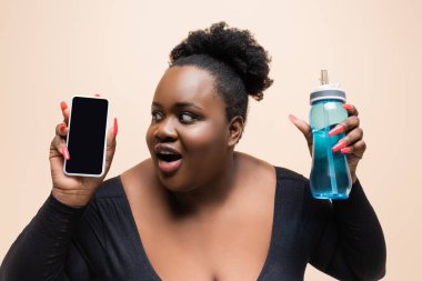 Şaşırmış Afro-Amerikalı, elinde spor şişesi ve beyaz ekranlı akıllı telefon tutan iri yarı kadın. 