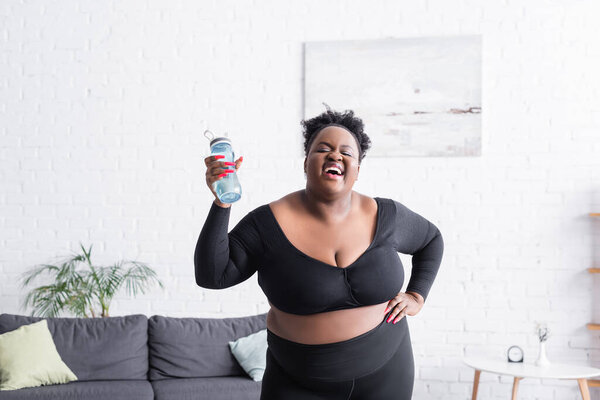 счастливый африканский американец плюс размер женщины в спортивной одежде стоя с рукой на бедре и держа спортивную бутылку 