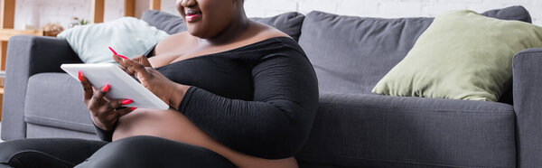 обрезанный вид на довольный африканский американец плюс размер женщины в спортивной одежде с помощью цифрового планшета в гостиной, баннер