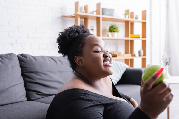 улыбается африканский американец плюс размер женщины в спортивной одежде держа зеленое яблоко 