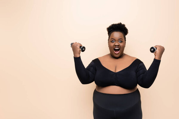 изумленный африканский американец плюс размер женщины в спортивной одежде держа гантели изолированы на бежевый 