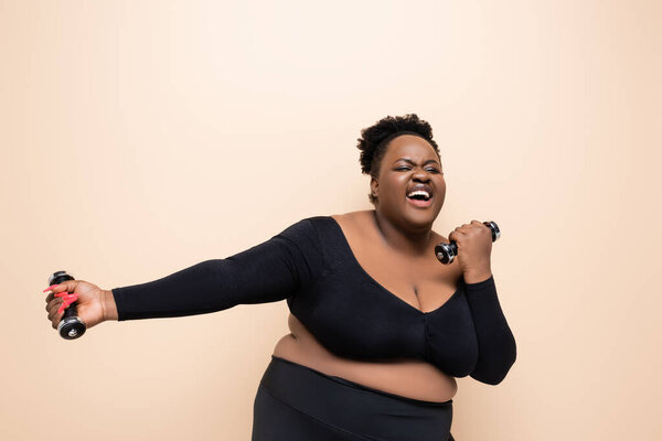 веселый африканский американец плюс размер женщины в спортивной одежде держа гантели изолированы на бежевый 