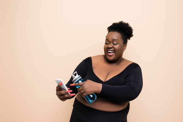 счастливый африканский американец плюс размер женщины в спортивной одежде держа спортивную бутылку и указывая пальцем на смартфон изолирован на бежевый 