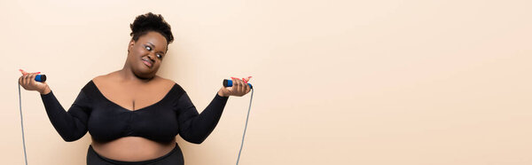 Африканский американец плюс размер женщины в спортивной одежде держа скакалку изолированы на бежевый, баннер