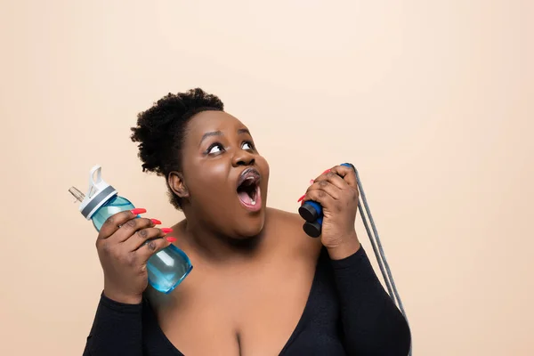 shocked african american oversized woman in sportswear holding sports bottle with water and skipping rope isolated on beige