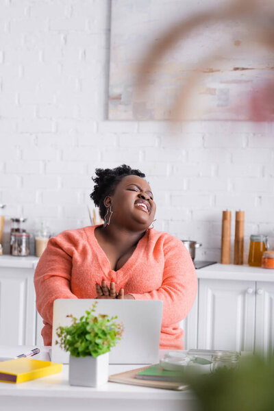 happy african american plus size freelancer using laptop and laughing at home