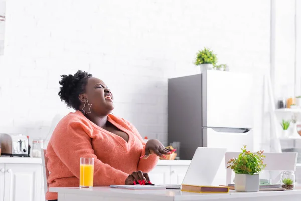 happy african american plus size freelancer laughing near laptop and orange juice on table
