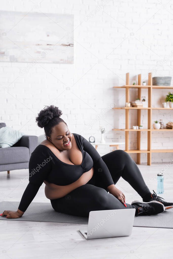 cheerful african american plus size woman watching online training on laptop while sitting on fitness mat 