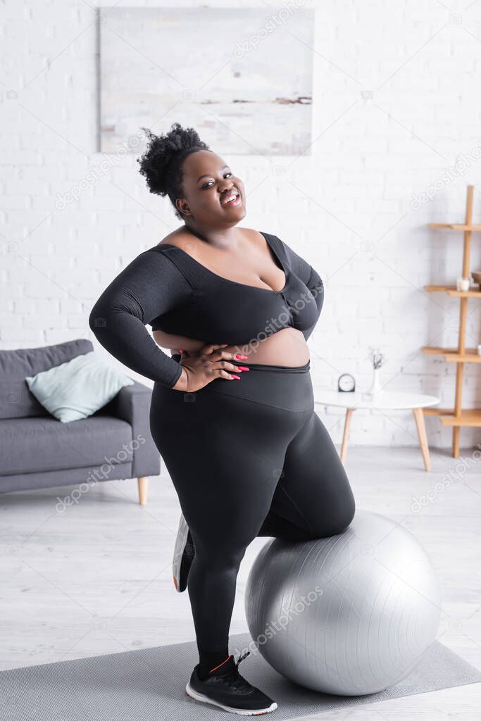 cheerful african american plus size woman in sportswear leaning on fitness ball