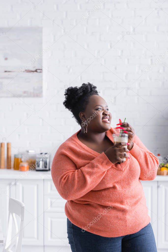 smiling african american plus size woman holding plastic cup with tasty smoothie in kitchen 