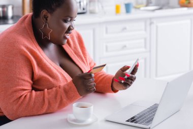 Mutlu Afro-Amerikan artı boyunda bir kadın kredi kartı ve akıllı telefon tutuyor.
