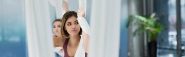 young sportswoman looking away while training with fly yoga hammock, blurred foreground, banner clipart