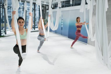 Genç sporcu kadınlar spor salonunda yoga hamaklarıyla ısınıyor.