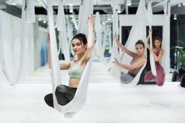 Nilüfer tatbikatı yaparken mutlu bir kadın kafasını çeviriyor. Arka planda bulanık yoga hamağında poz veriyor.