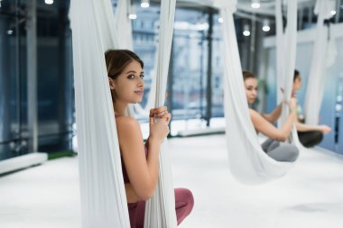 young woman looking away while sitting in hammock in lotus pose on blurred background clipart