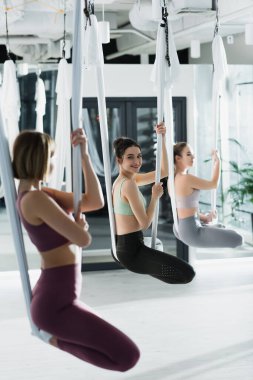 young sportswomen practicing lotus pose in aerial yoga hammocks, blurred foreground clipart
