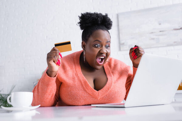 amazed african american plus size woman holding credit card and looking at laptop 