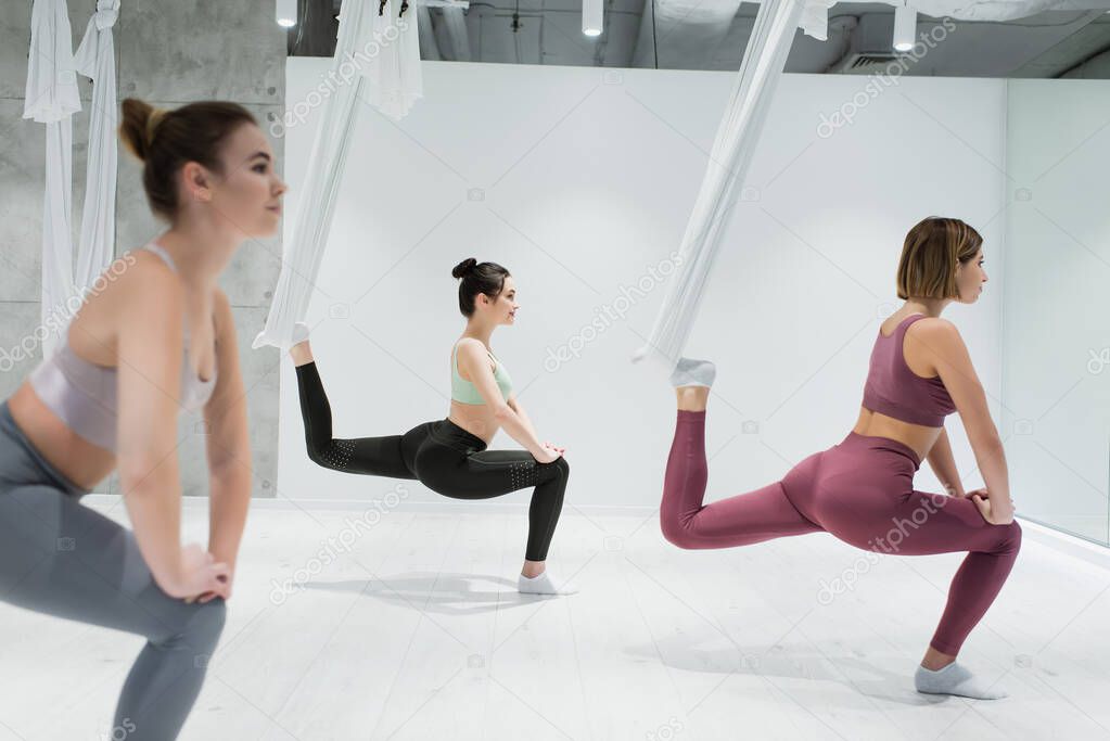 sportive women stretching with fly yoga hammocks in fitness center, blurred foreground
