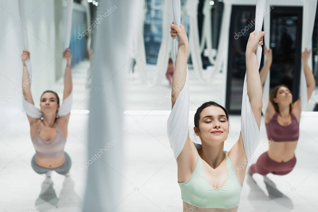 group of young sportswomen stretching with aerial yoga straps on blurred foreground