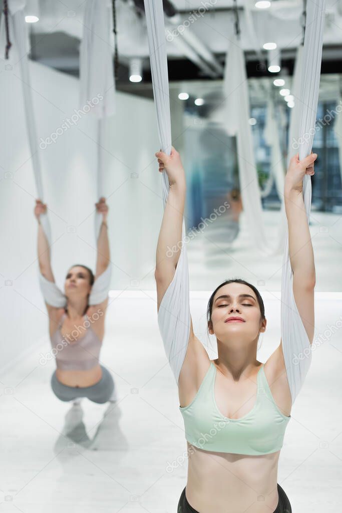 young woman with closed eyes training with aerial yoga strap on blurred background