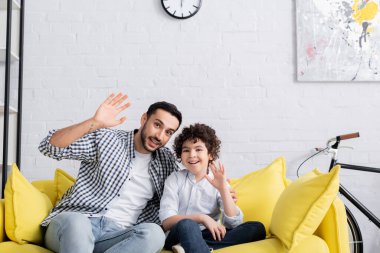 Neşeli Müslüman baba ve oğul koltukta otururken kameraya el sallıyorlar.