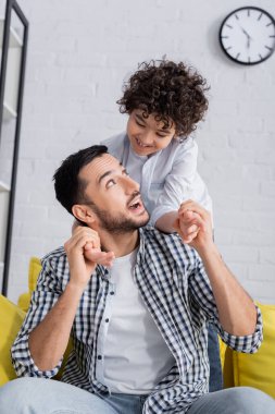 excited arabian man holding hands of happy son while having fun at home clipart