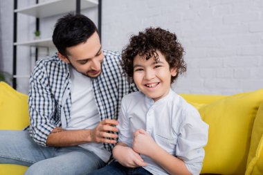 cheerful arabian man tickling joyful son while sitting on sofa at home clipart