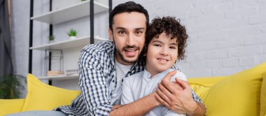 Neşeli Müslüman adam kameraya, afişe bakarken mutlu oğlunu kucaklıyor.