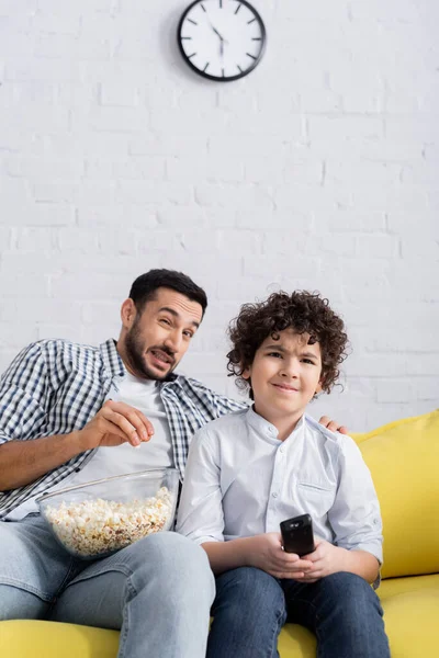 frightened arabian man eating popcorn while watching tv with son at home