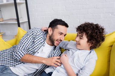 excited muslim man tickling happy son while having fun on sofa clipart