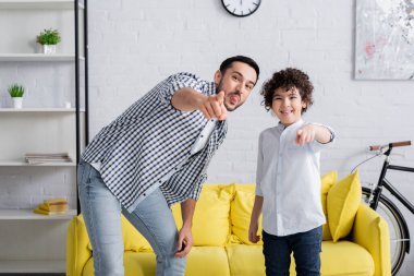 cheerful arabian man and son sticking tongues and pointing with fingers while looking at camera clipart