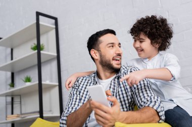 Mutlu Müslüman çocuk gülümseyen babasının ellerinde cep telefonunu işaret ediyor.