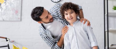 Mutlu oğlunun omuzlarına sarılan neşeli Arap adam kameraya bakıyor, afiş.