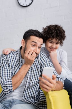 excited arabian dad and son looking at mobile phone at home clipart