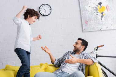 Neşeli Arap adam heyecanlı oğlunu işaret ediyor. Yumruklarını sıkarak kanepeye zıplıyor.