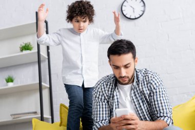 hoşnutsuz Arap çocuk, babasının yanında el kaldırmış akıllı telefondan mesaj atıyor.